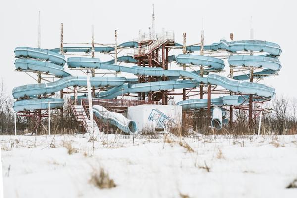 A large, deteriorating water slide complex photographed in winter, with multiple intertwining blue slides winding around a tall central structure. The slides appear abandoned and unused, set against a snowy landscape with bare trees in the background. The infrastructure looks weathered and in disrepair, creating a stark, desolate atmosphere.