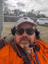 A picture of a white man with a grey and brown beard. He is wearing a fawn coloured hat with a HRCCQ logo, orange hi-viz shirt and black sunglasses. He has a radio headset on.