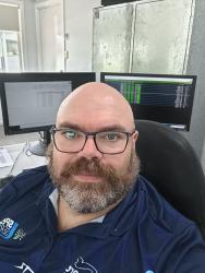 A white, balding man with a grey and brown beard. He is wearing a blue polo shirt and glasses, sitting in front of computer screens showing timing information. He looks very official!