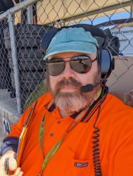 A picture of a white, balding man with a grey and brown beard. He is wearing a dull-green GitHub cap with a radio headset over it, dark sunglasses, a bright orange hi-vis short and a lanyard around his neck holding a whistle. He looks very official!