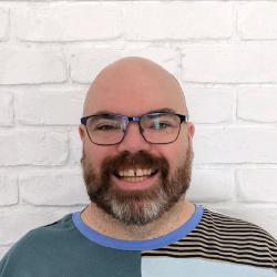 A picture of a white, bald man with a grey and dark brown beard. He is wearing a patchwork-style t-shirt and black rimmed glasses.